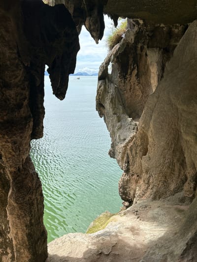 The image is a vertical, eye-level shot taken from within a cave, looking out towards the ocean. The foreground is dominated by the rough, textured surfaces of the cave walls, which are dark and cast shadows. On the left, a large, dark, stalactite-like formation hangs down. On the right, a lighter, more jagged rock face curves into the frame.
Through the opening of the cave, a body of calm, green water is visible, with gentle ripples on its surface. In the distance, a small boat with a single sail is visible on the water. The horizon shows a hazy outline of islands or landmasses under a bright, slightly cloudy sky. The overall impression is one of a natural, secluded viewpoint overlooking a serene seascape.
This scene is characteristic of the limestone karst landscape found in areas such as Phang Nga Bay, near Krasom, Thailand. Such caves and viewpoints are common attractions in this region, offering dramatic perspectives of the emerald waters and scattered islands. The cave provides a natural frame for the ocean view, highlighting the contrast between the dark, enclosed space and the bright, open expanse of water and sky.