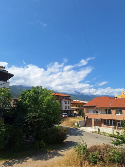 This image depicts a sunny residential scene in Petrich, Bulgaria, looking towards a prominent mountain range. The setting is a quiet neighborhood, likely situated on a gentle slope, with houses built into the landscape.

The upper half of the image showcases a clear, bright blue sky filled with numerous white, fluffy cumulus clouds. These clouds are particularly dense along the horizon, where they meet the distant mountains. The mountains, which form a significant backdrop, are partly obscured by the clouds and atmospheric haze, but their green, forested slopes are discernible.

In the midground and foreground, several multi-story residential buildings are visible. Most of these houses feature distinct reddish-orange tiled roofs and walls painted in warm, earthy tones such as brown and orange-yellow. Windows are clearly visible on their facades. A large, verdant tree with dense green foliage stands prominently in the mid-ground, and other green shrubs and bushes line a dirt or unpaved path that curves through the scene on the right side. This path is bordered by dry, yellowish grass and weeds, suggesting a sunny, possibly dry, time of year. A thin, dark utility wire stretches across the upper left portion of the sky. A white car is partially visible behind some greenery further down the path on the right.

The strong, distinct shadows cast by the houses and vegetation indicate that it is daytime, likely mid-morning or afternoon, under bright and sunny weather conditions. There are no people visible, and no discernible text appears in the image, contributing to a tranquil and serene atmosphere.