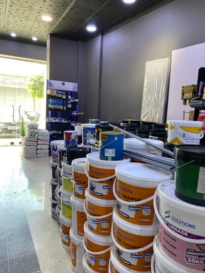 This is a medium shot, taken from a slightly low angle, of a retail store's interior in Baraki, Algeria. The store displays a large quantity of paint and construction materials. Dominating the foreground and midground are stacks of buckets and bags of what appear to be paint or plaster, neatly arranged in rows. The buckets are predominantly white and orange, with some black and green ones visible. Many of the buckets feature branding with the word "SOLUTIONS" and text in both English and Arabic. Some bags are stacked to the left of the scene, also bearing the "SOLUTIONS" logo.
In the background, a dark gray wall extends upwards, meeting a black ceiling with a textured, star-like pattern and recessed lighting. To the far left, through a large glass storefront, the exterior is visible, showing a street with parked cars and trees. One car has a visible license plate starting with "SX 83661". A display rack with various painting tools, such as rollers and brushes, is mounted on the back wall. The floor is a light-colored, speckled terrazzo. No people are visible in the image. The overall impression is one of a well-stocked retail environment specializing in building and decorating supplies.