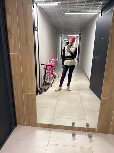 A person with a pink head covering is standing in a hallway holding a mobile phone. The hallway has a bike leaning against a wall, with doors on one side and a tiled wall on the other. The image appears to be a reflection in a mirror. The location is indeterminate, and the details of the scene do not provide evidence to confirm Verkhnii Liuk, Russia.