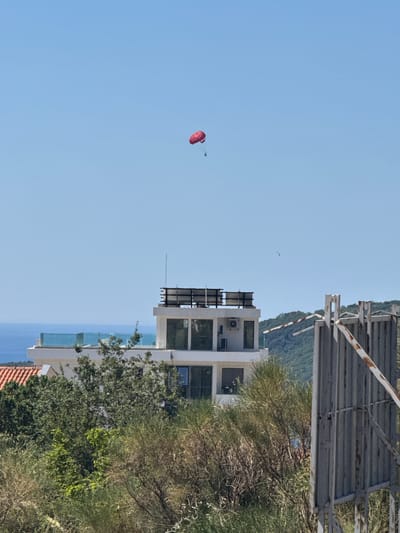 A red parasail with a person attached is visible in the sky above a modern white building with a flat roof and glass railings. The building is situated on a hillside overlooking the Adriatic Sea, with lush green vegetation in the foreground and on the slopes. A metal gate is partially visible on the right side of the image. The scene appears to be in a coastal town in Ivanovići, Montenegro, likely during the daytime given the bright sky and clear visibility.