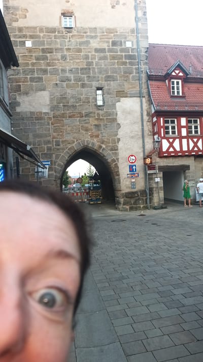 The image shows a street scene in Altdorf bei Nürnberg, Germany, in front of a historic stone gate. In the foreground, a person's eye is prominently featured, out of focus. Beyond the eye, the stone archway of the gate is visible, with buildings on either side. One building to the right has a red tiled roof and white timber framing. Several traffic signs are mounted on the wall of the gate, including a "2.8m" height restriction sign and directional arrows. A light-colored paved street is visible in front of the gate, and beyond the archway, construction equipment and barriers suggest ongoing work. Two people are visible standing in the doorway of the building to the right, and other individuals are faintly visible further inside the archway. The scene appears to be an outdoor public space.