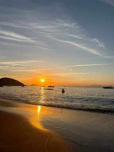 The image depicts a serene beach scene at sunset in Armação Dos Búzios, Brazil. The sun, a bright orange orb, is low on the horizon, casting a warm glow across the sky and water. Wispy clouds streak across a gradient of blue and orange sky. Gentle waves lap at the sandy shore, reflecting the sunset's golden light.
In the water, a lone individual is partially submerged, appearing to swim or relax. Several small boats are anchored in the calm sea. In the distance, a dark, silhouetted island rises from the water, with smaller islands and mountains visible further out. A sleek, white motorboat is anchored to the right of the frame. The foreground is dominated by the wet sand of the beach, glistening with the reflection of the setting sun. The overall atmosphere is peaceful and tranquil.