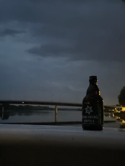 In Charnay-lès-Mâcon, France, at twilight, a brown glass bottle of Carlsberg Urpils beer stands on a dark, textured surface, likely a table or railing, in the foreground. The bottle is partially illuminated, with a label displaying the Carlsberg logo and text. Behind the bottle, a body of water, possibly a river, reflects the dim sky. A dark, horizontal structure, resembling a bridge or pier, spans across the water in the mid-ground. The background features a silhouette of trees and distant lights from buildings on the far bank. The sky above is overcast with dark, heavy clouds, suggesting impending rain or a storm. The overall atmosphere is moody and somber, with low light conditions and a muted color palette.