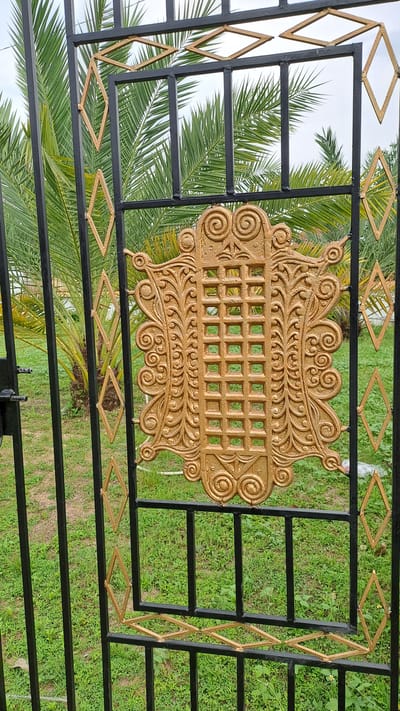 The image shows a close-up of a black metal gate with gold decorative elements. The gate features a central ornate gold medallion with intricate scrolling patterns and a grid-like design. In the background, palm fronds are visible against a light sky, suggesting an outdoor setting. The ground beneath the gate is covered in green grass. The overall impression is of a decorative entrance to a property. There are no people or specific actions depicted in the image. The location is identified as Meunasahteungkudigadong, Indonesia.