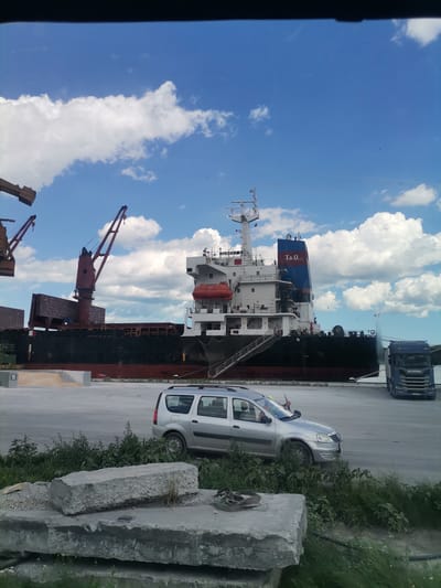 The image depicts a harbor scene in Ezerovo, Bulgaria, under a bright blue sky with scattered white clouds. A large cargo ship is docked, with cranes positioned nearby. In the foreground, a silver minivan is parked on a paved area. There are stacks of concrete blocks and lush green vegetation in the immediate foreground. A blue Scania truck is parked behind the minivan, to the right of the ship. The scene suggests ongoing port activity with the presence of heavy machinery and vehicles.