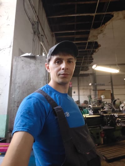 The image shows a medium shot of a man in a blue t-shirt and a black baseball cap, looking towards the camera. He has a strap from a bag or overalls slung across his chest. The man is in what appears to be a workshop or industrial setting. Behind him, there is a large, metallic cabinet on the left and several pieces of machinery, likely lathes, in the background. A fluorescent light fixture hangs from the ceiling, illuminating the space. The ceiling itself shows signs of damage and exposed beams. The overall environment suggests a utilitarian and possibly aged workspace. The context provided indicates this scene is located in Goczałków, Poland, a town known for its industrial presence, particularly in metalworking and manufacturing.