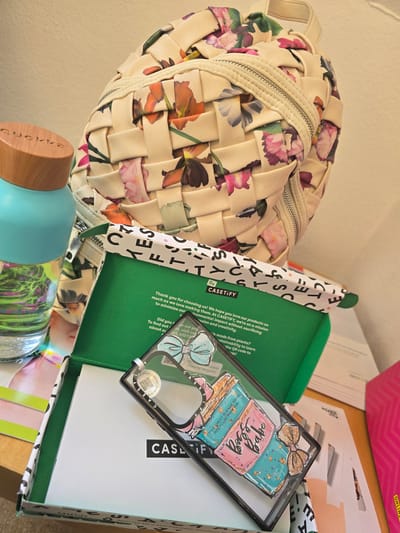 The image shows a floral woven backpack resting on a table or shelf. Next to the backpack is an open Casetify box. Inside the box, there is a green card, a white box, and a Casetify phone case with a "Dog Babe" design. A water bottle with a wooden cap is at the bottom of the image. The location is not evident from the image.