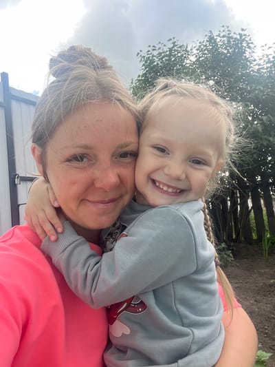 A close-up selfie of a woman and a young girl embraces outdoors. The woman, with her hair tied up in a bun, wears a bright pink sweatshirt. The girl, with blonde hair in braids, is wearing a light blue sweatshirt and is hugging the woman. They are both smiling. In the background, there is a fence, trees, and a cloudy sky. The image appears to be taken in a residential area, possibly a backyard. The location is identified as Sukhinskoe, Russia, suggesting a rural or suburban setting.