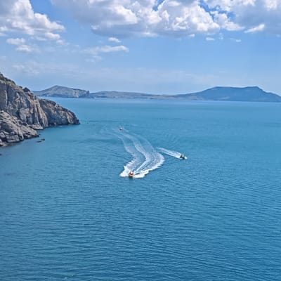 The image depicts a view of the Black Sea from a cliffside overlooking the water. A rocky coastline with sparse vegetation is visible on the left side of the frame. Several small boats are out on the water, leaving white wakes behind them. In the distance, rolling hills and mountains form a hazy horizon under a blue sky with scattered white clouds. The overall scene suggests a recreational activity on a clear day, likely in a coastal region. The location is the city of Sudak, Ukraine.