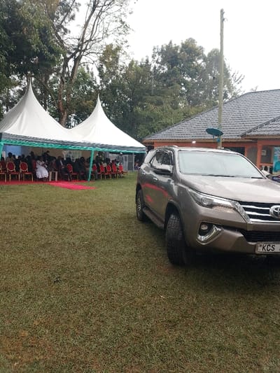 This image depicts an outdoor gathering in the Marenyo-Shianda ward, Kenya. The scene features a grassy area under an overcast sky, with dense green trees in the background.
In the foreground, a brown Toyota Fortuner SUV, identifiable by the initial characters "KCS" on its license plate, is parked on the grass, facing left. To the left of the vehicle, two white, peaked canopy tents, accented with light blue/green trim, are erected. Beneath these tents, a group of individuals is seated on red chairs arranged in rows, facing towards the right. A red carpet or runner is laid on the grass in front of the seated individuals. Most attendees are dressed in dark clothing, with one individual prominently visible in a white dress.
To the right of the SUV, a building with a dark-tiled roof and an orange/brown painted wall is visible. A small satellite dish is affixed to the side of this building, near a tall wooden utility pole. A partially visible poster or banner, displaying a person's image and text, is affixed to the building's wall. The individuals under the tent appear to be attending an organized event or function.