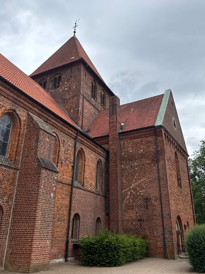 The image shows the exterior of the St. Marien Church in Bassum, Germany. The church is constructed of red brick, with a steeply pitched red tile roof. Several arched windows are visible on the brick facade. A tall, square tower with a pointed roof topped by a weather vane rises from the center of the church. Lush green bushes are planted in front of the building, and a gravel path leads to an entrance. The sky above is overcast.