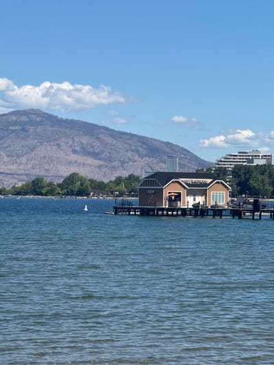 The image depicts a wide shot of Okanagan Lake in Kelowna, Canada, on a clear, sunny day with scattered clouds. In the foreground, the blue water of the lake ripples gently. A small, sandy beach area is visible at the bottom edge of the frame. In the middle ground, a brown and tan boathouse with a black roof sits on a wooden pier extending into the lake. The boathouse has "THE BOATHOUSE" and "CREW" written on it. Several dark-colored boats are tied to the pier. A white buoy is floating in the water near the boathouse.
In the background, a line of green trees and buildings forms the shoreline. Beyond the trees, two modern, multi-story buildings are visible, one white and one with horizontal lines. A large, barren mountain looms in the distance, its slopes appearing somewhat scorched or dried out, with sparse vegetation. The sky above is a vibrant blue. The overall scene is serene and suggests a popular waterfront area.
