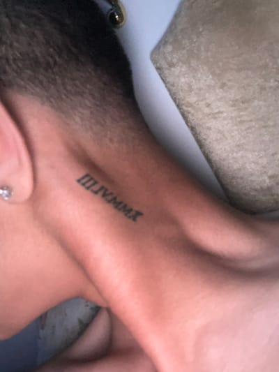 The image shows a close-up of a person's neck with a tattoo of Roman numerals "IIIIVMMX" on the back of their neck. A small, sparkly earring is visible on the person's earlobe. The background consists of a textured, beige surface and a lighter, possibly white surface. The scene is a close-up shot, and the location in Ognianovo, Bulgaria, is not visually discernible from the image itself.
