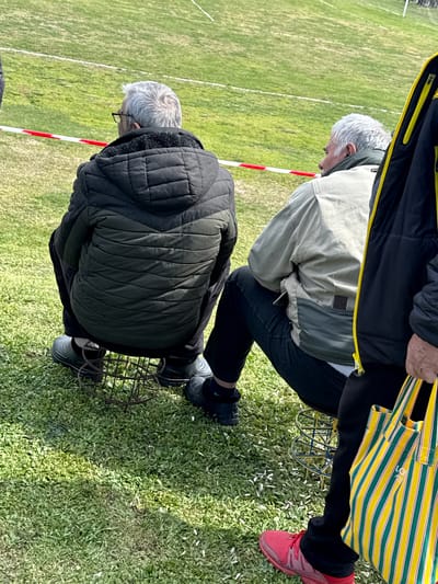 The image portrays an outdoor scene in Petarnitsa, Bulgaria, with three adult males observing an area beyond a red and white striped barrier tape. Two men are seated with their backs towards the viewer on cylindrical metal grates, positioned on a grassy field. The man on the left has gray hair, wears glasses, a dark gray padded jacket with a hood, dark pants, and dark shoes, and sits on a rusty, dark-colored metal grate. To his right, a second man with gray hair wears a light tan jacket over dark pants, seated on a similar, partially visible metal grate. The background displays a broader grassy field marked with faint white lines, characteristic of a sports ground. A third individual is partially visible on the far right, wearing a black jacket with yellow trim, dark pants, and red sneakers, holding a yellow and green striped tote bag in his right hand. The immediate ground is covered in green grass with scattered small white debris.