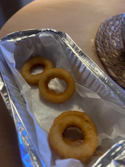 A close-up, overhead shot shows four golden-brown onion rings arranged in a foil container lined with white paper. Three rings are clustered near the top, with two overlapping, while the fourth is positioned below them. The rings have a crispy, battered exterior. The container sits on a light-colored, wooden surface, and a woven placemat is visible in the upper right corner. The lighting suggests an indoor setting. The image is taken in Yastrebovskii, Russia.