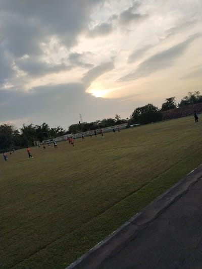 The image depicts an outdoor soccer field in Botok, Indonesia, during what appears to be late afternoon or early evening. The sky is overcast with dramatic clouds, and the setting sun is visible as a bright glow behind a break in the clouds, casting a warm light over the scene.
There are approximately 15-20 individuals on the field, most of whom appear to be young males engaged in a soccer game or practice. They are dressed in various colored jerseys and shorts, some in orange, some in blue, and others in dark green. Several individuals are in motion, running across the grassy field, suggesting active play. One person in a black uniform stands near the right edge of the frame, seemingly observing the game.
The field is large and covered in well-maintained green grass. A white line is visible marking the edge of the field. In the background, there is a brick wall with a white painted section that has some text on it, possibly a stadium name or sponsor advertisement. Beyond the wall, there are trees and vegetation, and a tall transmission tower is visible in the distance. The foreground shows a portion of an asphalt path or road bordering the grassy field. The overall impression is a casual sporting event taking place in a semi-rural or suburban setting.