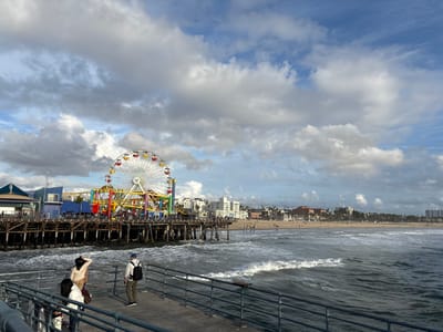 Santa Monica Pier is captured on a partly cloudy day, with the Ferris wheel prominently displayed amidst colorful structures. A group of people stands on the pier's edge, overlooking the ocean and the sandy beach. Waves gently crash against the shoreline, and buildings line the horizon. The sky is filled with puffy clouds, creating a scenic coastal view.