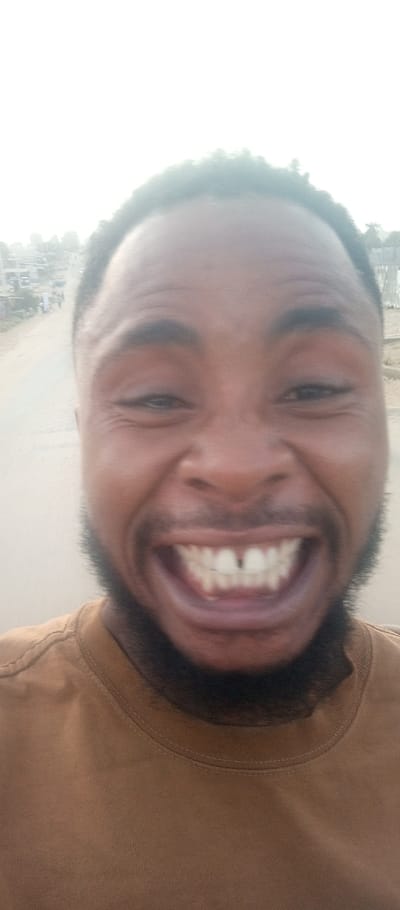 A close-up selfie shows a Black man grinning widely, displaying his teeth, including a noticeable gap between his upper front teeth. He has short dark hair and a full beard. He is wearing a brown t-shirt. A blurred background suggests he is outdoors, possibly on a road in a residential area. The light is bright and somewhat diffused.