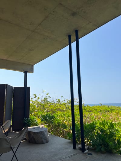The image shows a view from an outdoor patio in San Isidro Llano Grande, Mexico (latitude: 15.939, longitude: -97.279). The patio is partially covered by a concrete roof supported by two black pillars. To the left of the pillars, there are two beige lounge chairs. To the right of the pillars, there is a wooden table with a piece of wood on top. The patio floor is made of gray concrete. There are green bushes and trees in the background. The sky is blue and clear. The ocean is visible in the distance, beyond the trees.