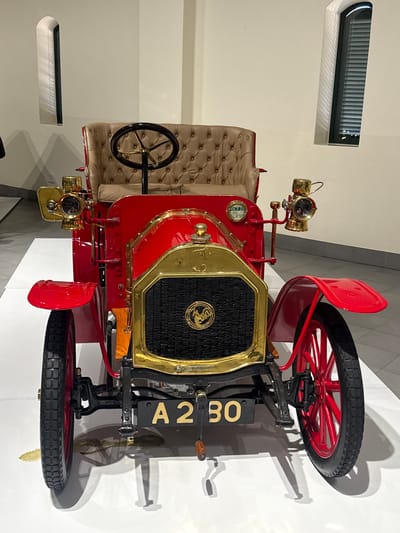 The image shows a vintage red and gold car with black tires and a black grill. The car is in a museum setting with white walls and a modern window fixture. The car's license plate reads "A 280." The photograph appears to be taken at the Rupert Museum located in Stellenbosch Local Municipality, South Africa.