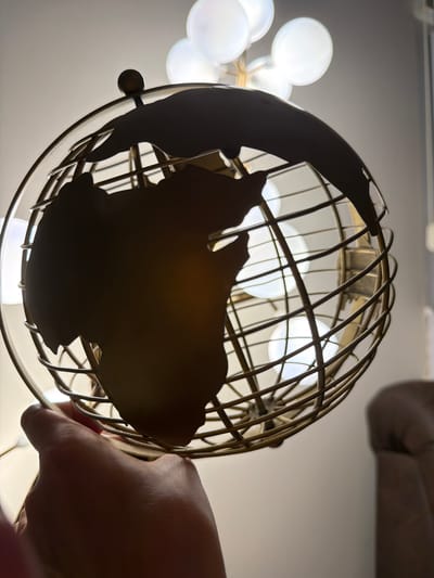 A hand holds a gold wire globe of Earth, obscuring much of the globe's continents, which are cutouts within the wireframe. The globe is lit from behind by several bright, diffused light bulbs, creating a soft glow through the wire structure and the cutouts. The background is a plain, light-colored wall. To the right, a blurred, upholstered piece of furniture, possibly a sofa, is visible. The scene is indoors. The image does not contain information indicating it is located in Mojanovići, Montenegro.
