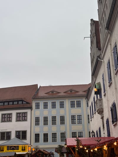 The image displays a daytime scene in the Markt (Market Square) of Meissen, Germany, under an overcast sky.
In the foreground right, a multi-story, light-colored building with a gabled roofline is visible. Its windows are fitted with dark blue shutters, three of which are numbered '21', '22', and '23', indicating an Advent calendar display. An ornate, carved stone balcony is positioned on the facade below one window. A gold, star-shaped light hangs from an archway or awning on this building. A metal downspout extends from the upper gabled section.
Adjacent to this, toward the left and center, are several other multi-story historical buildings. One building is light blue with cream-colored rectangular panels surrounding its dark-framed windows. Its reddish-brown tiled roof features two distinct oval "eyelid" dormer windows. To the far left, another off-white building with dark-framed windows also has a reddish-brown tiled roof with a dormer window. Partially visible text on its facade includes "MEISSEN" and "Schwalbennest."
The lower portion of the image shows several temporary market stalls. One on the far left has a yellow canopy with "Schwalbennest" text and string lights. Adjacent stalls feature dark canopies and are decorated with string lights and evergreen foliage. A sign with "Weltladen" is partially visible behind one stall. No individuals are discernible within the frame.