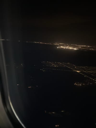 This is an image of city lights observed at night from an aircraft, potentially taken from the air looking toward the city of Kastrup, Denmark. The lights appear as streaks and blurs due to the speed and motion of the aircraft or possibly the camera settings. The darkness surrounding the lights suggests a night sky and the absence of other sources of light. A portion of the aircraft's window is visible in the lower part of the image. The lights are primarily concentrated on the left side of the image and along some linear patterns to the right, suggesting roads or developed areas.