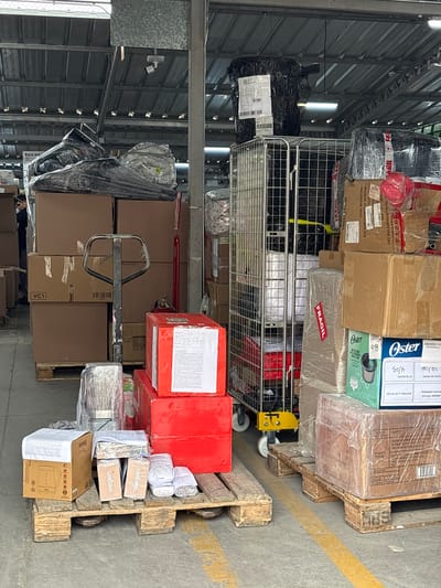 This image shows a storage or warehouse area with many stacked boxes and packages. In the foreground, there's a pallet with several boxes and some smaller items. One of the boxes is red and has white text on it. There are also some smaller cardboard boxes labeled "siegen." A pallet jack is positioned in the middle of the image, resting on top of some stacked brown boxes. The scene is indoors, likely in a warehouse, with metal shelving units visible in the background. The lighting suggests it might be daytime.

There are several labels with text visible on the packages. Some of these include "PACK-CRYGUARENAS-CRY," "MEDIDAS: 47.5CMX34CMX54.5CM," "PROVEEDOR: ANNA," and "HECKC CHINA." One box has the brand name "Oster" and mentions "FRAGIL" (fragile). Other text includes "Dinet," "HERVIDOR," and "THOMAS." Some labels indicate shipping information with dates like "19/06" and "4/05" along with "ORIGEN: TOTTUS PUNTA HERMOSA" and "DESTINO: LI 3P CD VES."

The overall impression is a busy logistics or storage environment where goods are being organized and prepared for transport or distribution. No people are clearly visible in the image, although a hint of a person can be seen at the bottom right of the frame near the boxes.