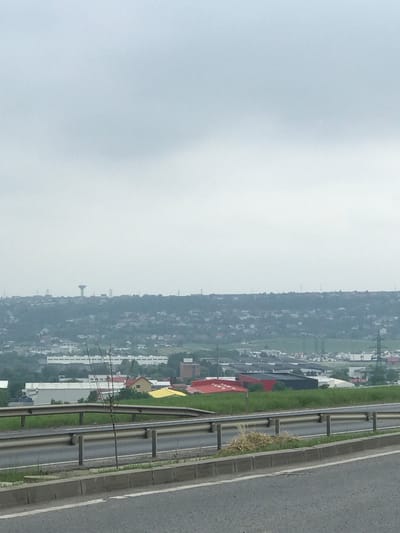 The image displays an elevated view of the urban and semi-urban landscape of Șcheia, Romania, under an overcast and hazy light-grey sky, which occupies the upper portion of the frame. The horizon reveals rolling terrain densely populated with residential and commercial structures. A prominent, mushroom-shaped water tower is visible on the distant skyline, slightly to the left of the image's center, accompanied by several smaller communication masts.

In the mid-ground, a mixture of buildings with various roof colors, including white, red, yellow, and dark grey/black, are interspersed with green patches of vegetation. Power line pylons are discernible on the right side, extending into the mid-distance.

The foreground consists of a section of a highway or major road. A grey asphalt surface with a white line delineates the road edge, followed by a concrete curb. Above the curb, a dual-level metal guardrail system runs horizontally across the frame. Behind the guardrail, a grassy median or shoulder contains a small, slender, leafless tree or shrub positioned slightly to the left of the center. Patches of dried grass are also visible in this area, particularly on the right side. No people or animals are present.