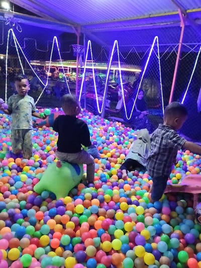 The image displays an indoor recreational ball pit facility located in Namplohbaro, Indonesia, observed at night. The play area is enclosed by a mesh fence and covered by a corrugated metal roof supported by metal beams. Blue neon strip lights, arranged in a zigzag pattern, provide the primary illumination, casting a distinct blue hue over the scene. A small basketball hoop with a white net is visible on the left side, attached to a support beam.
Thousands of multicolored plastic balls fill the entire floor of the enclosure. Four children are present within the ball pit. On the left, a young male child, approximately 4-6 years old, with short dark hair, stands facing forward, holding two colored plastic balls. In the center, another young male child, of similar age, is viewed from behind, sitting on a lime-green, animal-shaped bouncy toy. To the right, a third young male child, also approximately 4-6 years old, is partially visible from behind, wearing a plaid shirt and seemingly interacting with the balls. A fourth child, wearing a light-colored hooded garment, is partially submerged in the balls in the midground, seen from the back.
In the background, outside the ball pit enclosure, an adult female wearing a dark headscarf (hijab) and a patterned long-sleeved top is seated, leaning against a red motorcycle, observing the children. Several other motorcycles are parked in the background, and indistinct figures are visible further behind the fence. The children are actively playing within the ball pit, while the adult appears to be supervising.