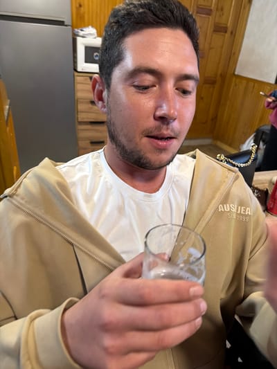 A man is holding a glass with some liquid in it. The man has dark hair, and a beard, and is wearing a white t-shirt and tan jacket. The background shows a kitchen with wood paneling, a refrigerator, and other kitchen appliances. There is no evidence to suggest the photo was taken in Lilkovo, Bulgaria.