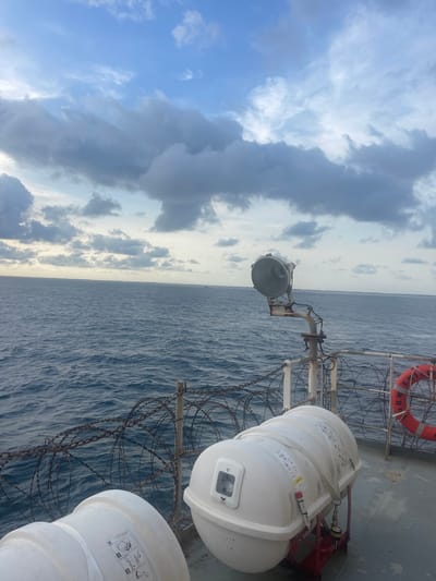 The image displays an exterior view from the deck of a vessel situated at sea, identified as being in the vicinity of Orimedu, Nigeria. The foreground features a grey ship deck. Two white, capsule-shaped marine life rafts are visible, secured within red launching cradles; one is prominently positioned in the lower right, displaying safety instruction decals. A red lifebuoy, partially inscribed with "ARMER...", hangs from a white metal railing on the right side of the deck. Coils of barbed wire are strung along the ship's perimeter railing, forming a security barrier. A large, circular searchlight is mounted on a rusty metal pole, angled over the water. The midground shows a wide expanse of deep blue-grey ocean water with moderate ripples, extending to a distant horizon. The background consists of a sky with varying shades of light blue interspersed with numerous grey, cumulus-type clouds. No individuals or specific actions are depicted.