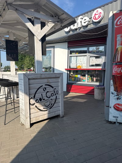 The image shows an outdoor cafe called "Cool Cafe" located in Ivatsevichy, Belarus. The cafe has a white wooden canopy with string lights, and a counter window with red trim. There are menus displayed on the window, and various snacks and drinks are visible inside the display case. A Coca-Cola vending machine is positioned to the right of the cafe. In the foreground, there is a wooden planter box with the "Cool Cafe" logo painted on it, and a bar stool at a wooden table. The ground is paved with interlocking bricks. The scene is well-lit by sunlight, casting shadows on the ground. The overall atmosphere appears to be casual and inviting, suitable for a quick refreshment.