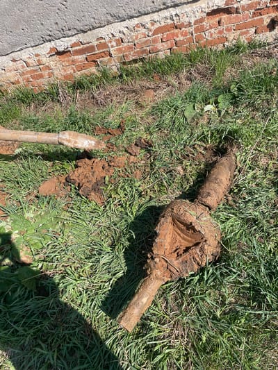 The image depicts a partially excavated pipe fixture coated in mud, resting on grass and dirt. A brick wall runs along the left edge of the frame, bordered by a section of concrete. A shovel is partially visible, also covered in dirt, near the excavated area. There are no people visible.