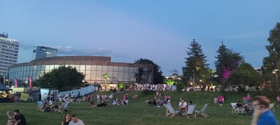 The image shows an outdoor evening gathering in Linz, Austria, likely at the Open Air Festival grounds near the Ars Electronica Center. A large, modern building with a curved glass facade is visible in the background. In the foreground, a grassy area is filled with people seated on blankets and in lounge chairs, enjoying the atmosphere. The sky is a deep twilight blue, with some clouds present. Several trees dot the landscape, and decorative lighting is visible, including some with a pink hue. Booths and tents suggest a festival or event is taking place. People are engaged in conversation, relaxing, and observing their surroundings. The overall scene conveys a sense of community and leisure.