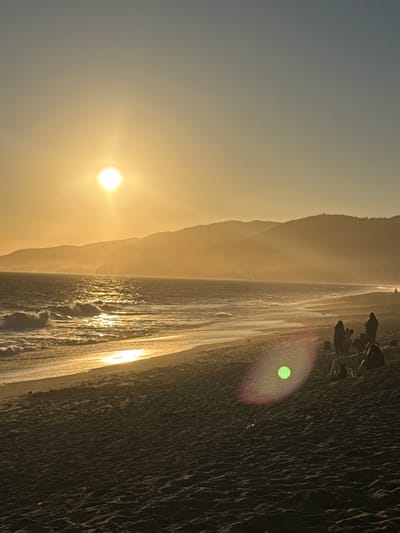 This is a vertical, medium-shot, eye-level, natural light photograph of a beach in Malibu, California, USA, during sunset. The sun, a bright orb, is positioned in the upper left quadrant of the frame, casting a golden glow over the scene. The sky transitions from a clear, pale blue at the top to a warm yellow-orange gradient near the horizon. Distant mountains form a silhouetted backdrop, shrouded in a light haze. The ocean is calm, with gentle waves breaking on the shore, their surfaces shimmering with the sun's reflection. The wet sand near the water's edge reflects the golden light, creating a bright band along the shoreline. Further up the beach, the dry sand is textured with small ripples.
In the middle ground, on the right side of the frame, a group of people are gathered. One person appears to be standing, while others are seated or reclining on the sand. A white dog stands near the seated figures. To the left of this group, a bright green lens flare with a pink halo is visible, also on the sand. The overall atmosphere is tranquil and serene, characteristic of a sunset on the California coast.