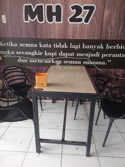 The image depicts a close-up view of a table and chairs in what appears to be an outdoor seating area of a cafe or restaurant. The main focus is a light-colored wooden table with a dark metal frame, set on a white tiled floor. Two black plastic chairs with a curved design are visible, one on the left and another partially on the right, with a third chair's leg visible in the background on the right.

On the table, there is an orange plastic holder, possibly for sugar packets or napkins, with the word "pulpios" printed on it. The table surface has some subtle markings and a darker circular outline of what might have been a coaster or an object.

In the background, a large black banner with white lettering is displayed. The prominent text at the top reads "MH 27". Below this, in smaller white text, is a quote in Indonesian: "Ketika semua kata tidak lagi banyak berbica maka secangkir kopi dapat menjadi perantara dan mencairkan semua suasana." This translates to "When words are no longer many, a cup of coffee can be an intermediary and melt all the atmosphere." The banner has a wood-grain texture and appears to be mounted on a wall.

The environment suggests a casual public space. The white tiled floor shows some minor debris, including a few cigarette butts scattered around the legs of the table. The lighting indicates it is daytime. The scene is static, with no people actively engaged in any actions. The overall impression is of an unkempt but functional seating area.