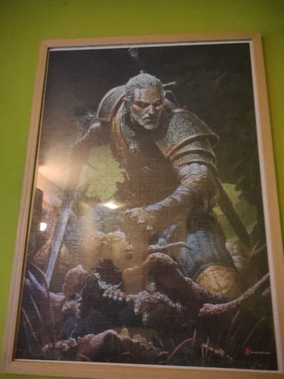 The image shows a framed jigsaw puzzle depicting a character from The Witcher, likely Geralt of Rivia. The character is a grizzled man with white hair and a scar across his face, wearing medieval-style armor and chainmail. He is holding a sword in his right hand. The background is dark and appears to be a forest or natural environment with some gnarled tree-like structures in the foreground. The puzzle is hanging on a wall painted a bright, lime green. There are reflections on the surface of the puzzle, indicating it is under some form of lighting. There is no discernible location in the city of Figueres, Spain, provided by the image.