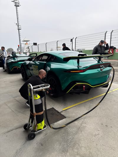 The image depicts a pit lane scene at a motorsport circuit, identified as the Circuito Tazio Nuvolari in Cervesina, Italy, under an overcast sky.

In the foreground, a bald, bearded man in a black shirt and dark pants is crouched beside the rear-left wheel of a green Aston Martin Vantage race car. He is operating a tire pressure adjustment system, with a black hose extending from a silver-framed, two-wheeled cart holding a black and yellow-green gas cylinder. The car features a large rear wing, a prominent rear diffuser, and yellow-green accent lines along its bodywork. Visible markings on the car include "LC.172", "958", and "SANTEK".

Behind this vehicle, a second identical green Aston Martin Vantage race car is parked with its driver's door open. A helmet rests on its roof. A man in a white racing suit with blue accents stands by this car. Further back, two other individuals are present: one in a dark hooded sweatshirt observing the scene, and another in a dark jacket and backward baseball cap standing behind the closer car, touching his cap.

The ground is concrete with yellow painted lines and includes a metal drain cover. In the background, a tall metal mesh safety fence separates the pit area from the track. Elements of track infrastructure are visible, including a tall metal tower on the left, a small white structure marked "PIT OUT" with red lights, and a red wall-mounted fire extinguisher. The track surface is visible beyond the fence.