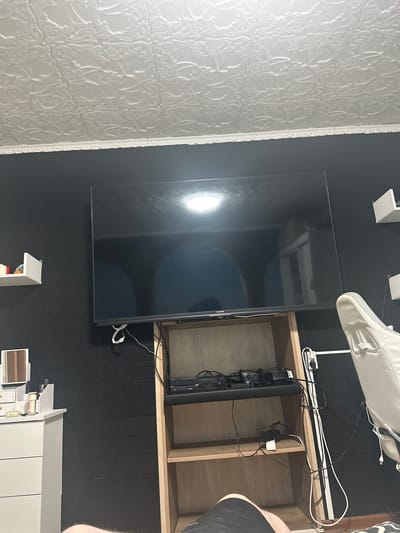 The image shows a television mounted on a black tiled wall in a room. To the right of the television is an open wooden shelf unit containing various electronic devices, including what appears to be a gaming console and cables. A white object, possibly a shoe or bag, is visible hanging from a mount above the shelf unit, with more cables connected to it. In the foreground, part of a white cabinet or furniture unit can be seen, with some items on top. The overall scene suggests a living space or entertainment area. The time of day and weather are not discernible from the image. There is no visible text in the image. The location context of Donskoy, Russia is provided but not visually evident within the image itself.
