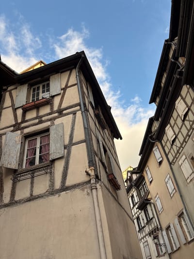 The image depicts a narrow street in Colmar, France, characterized by traditional half-timbered buildings. The perspective is low, looking upwards towards the sky which is blue with scattered white clouds. The buildings are a prominent feature, showcasing their distinctive wooden beams against a lighter colored plaster.

On the left, a building with pale beige walls and dark brown timber framing is angled towards the viewer. It features several windows. The lower window has red checkered curtains visible behind it and its light-colored shutters are open. Above it, another window with similar shutters is also visible, along with a small planter box. A dark metal pipe runs vertically along the side of this building, with a visible rusty joint.

To the right, another half-timbered building is visible, also angled, with its facade displaying a regular pattern of wooden beams and lighter colored infill. This building has multiple windows, most of which are closed with white shutters. The roofs of these buildings are dark, with visible gutters.

The overall impression is of a charming, historic European street, likely in the old town of Colmar, with architecture typical of the Alsace region. No people or animals are visible in the image. The lighting suggests it is daytime, possibly late afternoon, given the angle of the light and the shadows.
