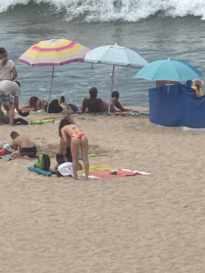 The image shows a crowded beach scene with numerous people and beach umbrellas. In the foreground, a woman in a red bikini is bent over. To her left, a man in dark swim trunks is seated on the sand. Several beach towels and bags are scattered around the foreground and middle ground. In the middle ground, a group of people are lying on the sand, some of whom are under beach umbrellas. The ocean with waves is visible in the background. The location appears to be a public beach.
