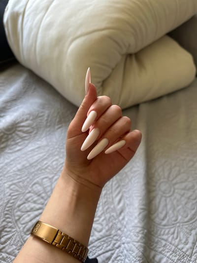 A person's left hand with long, almond-shaped, pale pink nails is resting on a textured, light grey blanket. The hand is positioned with the palm facing downwards and fingers slightly curled. The wrist is adorned with a gold-colored metal watch with a link band. In the background, a large, off-white, puffy pillow is visible, resting on the blanket. The scene appears to be indoors, likely in a bedroom. There is no indication of the city of Vorniceni, Moldova, in the image.