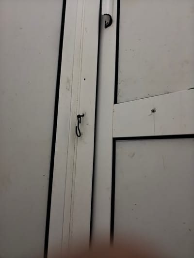 The image shows a close-up view of a white surface, likely part of a door or window frame. Two vertical white panels are visible, separated by a dark gap. One panel has a black wire or string attached to it, forming a loop or hook shape near the top. There are also some small dark specks and faint lines on the white surfaces, suggesting age or wear. The lighting appears to be from above and slightly to the right. No people are visible, and the scene is static. The image is an indoor shot, possibly in a residential or industrial setting. No text is visible. The location context provided is Ново-Талицы, Russia, but there are no specific visual cues within the image to confirm this.