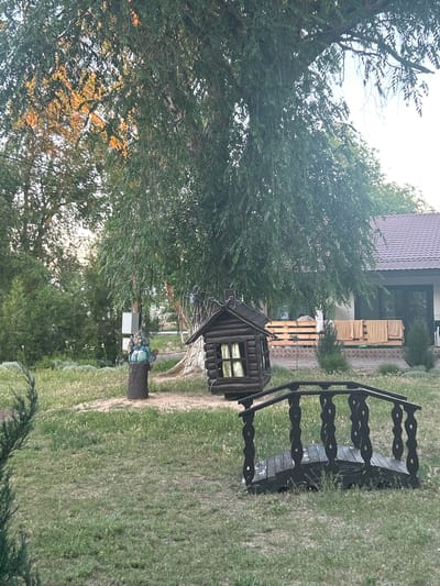 The image depicts a backyard scene in Останинское сельское поселение, Ukraine, featuring a variety of decorative and functional elements. In the foreground, a dark wooden footbridge with intricate railings arches over a patch of grass, suggesting a whimsical garden design. To the left of the bridge and slightly further back, a small, dark wooden birdhouse or decorative shed with a window is visible. A carved wooden figure, possibly a gnome, stands on a tree stump to the left of the birdhouse. In the background, a modern house with a tiled roof and large windows is partially obscured by the branches of a large, leafy tree. Several towels are draped over a wooden railing on the porch of the house, and the surrounding area is covered in grass and various types of bushes and trees, indicating a well-maintained outdoor space. The lighting suggests it is either early morning or late afternoon, with sunlight filtering through the leaves.