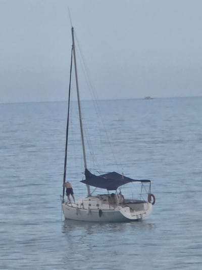 A sailboat is adrift in calm, light blue water, likely the Mediterranean Sea. The vessel is white with a single mast and a dark blue sail that is partially furled or damaged. A person is on board, appearing to be engaged in some activity near the stern. The scene is consistent with an open-water location in the vicinity of Sant Andreu de Llavaneres, Spain.