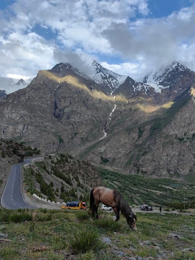 A horse grazes on a grassy slope in the foreground, its mane brushing the ground. Behind it, a winding mountain road snakes its way through rugged terrain. Several vehicles are visible on the road, including a tow truck carrying a blue car, a white car, and a silver car with people gathered around it. In the distance, towering, snow-capped mountains rise against a cloudy sky, with patches of sunlight illuminating their rocky slopes. The scene appears to be set in a mountainous region during the day, likely in a location with dramatic landscapes. The weather seems to be a mix of sun and clouds, creating dynamic lighting effects on the mountains. The overall impression is one of a serene, natural landscape with human activity interspersed.