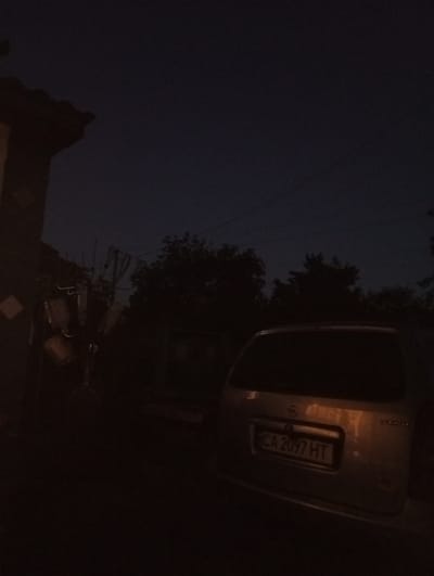 The image depicts a dark, outdoor scene at dusk or night, likely in Voznesenske, Ukraine. The primary subject is the rear of a silver minivan, illuminated by a warm, artificial light source. The vehicle has a license plate that reads "CA 2097 HT". To the left of the vehicle, a portion of a building or structure with a tiled roof is visible, along with some hanging objects that are difficult to discern due to the low light conditions. Trees and foliage are silhouetted against the darkening sky in the background, suggesting a semi-rural or residential setting. The overall mood is quiet and still, with no activity clearly indicated. The sky is a deep, dark blue, characteristic of twilight or nighttime.