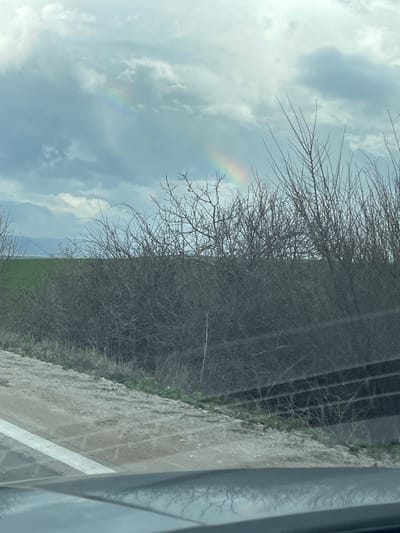 The image shows a view through a vehicle window in Belomortsi, Bulgaria. Outside, the sky is cloudy with a faint rainbow visible. A bare tree stands in the foreground, obscuring some of the view. Adjacent to the tree is a fence or barrier. The ground consists of a dirt or gravel path. The image is taken at an angle, with the right side of the frame showing part of the vehicle's interior.